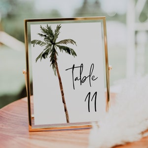 May include: A gold-framed table number card with a watercolor illustration of a palm tree. The card reads "Table 11".