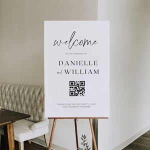 Op de afbeelding: Een wit rechthoekig welkomstbord voor een bruiloft op een houten ezel. Het bord bevat zwarte tekst met welcome, TO THE WEDDING OF, DANIELLE and WILLIAM, een zwarte QR-code en de tekst PLEASE SCAN THIS QR CODE TO VIEW OUR CEREMONY PROGRAM!