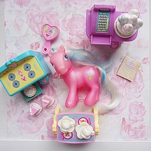 May include: A pink toy pony with a blue and white mane and tail, and a small ice cream cone graphic on its side. The pony is surrounded by toy accessories, including a stove, a cash register, and a menu with the text "Today's Specials".