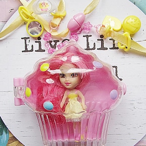 May include: A pink cupcake-shaped charm with a clear plastic lid. Inside the charm is a small doll with pink hair and a yellow dress. The charm is part of a necklace with a yellow ribbon and a variety of colorful beads.