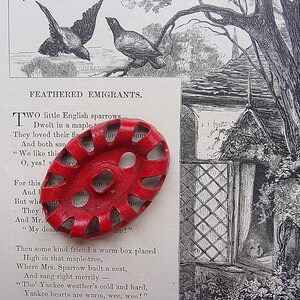 May include: A bright red, oval-shaped object with a series of holes, resting on a page of text. The text includes the title "FEATHERED EMIGRANTS." The background features a black and white illustration of birds and a cottage.