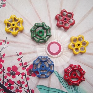 May include: A collection of colorful, flower-shaped valve handles in yellow, green, red, and blue. The handles are arranged on a decorative background with a floral design, showcasing a vintage aesthetic. These handles are likely for plumbing or industrial use.