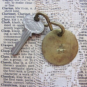 May include: Vintage key and brass tag with a loop, resting on a dictionary page with lace trim. The key is silver with the text "P&F Corbin" and the tag has an embossed design. The background is a collage of text and lace.