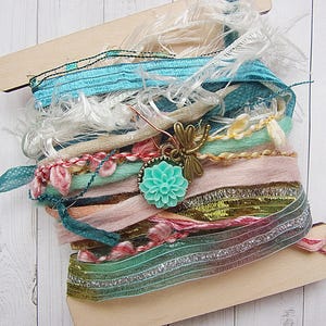 May include: A wooden spool of colorful ribbons and trims, including teal, pink, white, and gold. The spool is decorated with a small, green flower charm and a dragonfly charm.