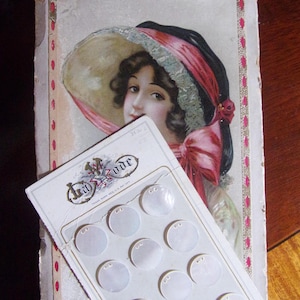 May include: A vintage card with a woman wearing a pink and white hat and a white dress. The card has a label that says "Hat-Vude" and features a display of 12 white mother-of-pearl buttons.