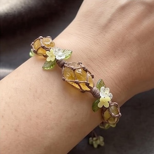May include: A macrame bracelet featuring raw yellow citrine stones encased in brown cord netting. The design is accented with small, pale yellow and translucent white flower-shaped beads and green leaf-shaped charms arranged along the cord.