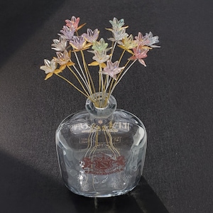 May include: A clear glass bottle with a decorative floral arrangement. The flowers are multi-colored, with pink, yellow, and green petals. The stems are gold-colored. The bottle has a round shape and a decorative emblem.