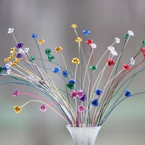 May include: A bouquet of colorful, wire-stemmed flowers in a small, silver vase. The flowers have metallic petals in various colors, including gold, red, blue, and green. The stems are thin and multi-colored, creating a vibrant display.