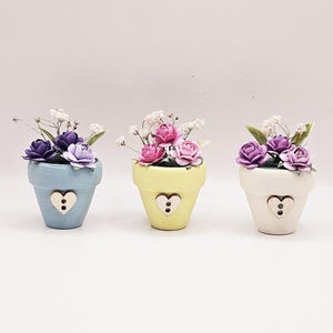 May include: Three small, decorative flower pots in pastel shades: light blue, yellow, and cream. Each pot has a heart-shaped cutout and is filled with colourful paper flowers and delicate white filler flowers. A charming, handcrafted floral arrangement.