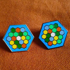 Puede incluir: Dos pines hexagonales de esmalte con un diseño de panal. Los pines son azules con hexágonos amarillos, verdes, naranjas y blancos.