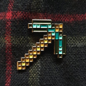 May include: A gold and blue pixelated pickaxe pin. The pickaxe is made of small squares and has a blue head and a gold handle.