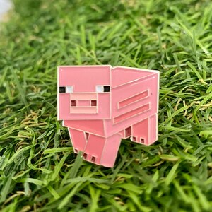 May include: A pink enamel pin in the shape of a pixelated pig. The pin has black and white eyes, a light pink snout, and a darker pink body. The pin is set against a background of green grass.