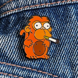 Peut inclure: Épinglette en émail orange représentant un singe fumant une cigarette. Le singe a un œil brun et un nez brun. Le singe tient un objet brun dans sa main gauche.