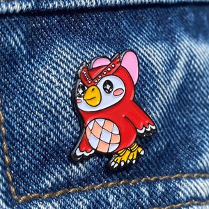 Peut inclure: Épinglette en émail représentant un hibou rouge et blanc, avec un contour noir. Le hibou a de grands yeux expressifs, un bec jaune et des oreilles roses. L'épinglette est posée sur un fond en denim bleu.