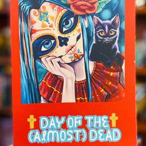 May include: A red journal with a colorful illustration of a sugar skull with blue hair and a black cat on its shoulder. The text on the cover reads "Day of the (Almost) Dead" and "A Meditation Guide/Journal for the 'Mature' Woman".