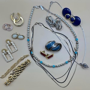 Puede incluir: Una colección de joyas vintage que incluye collares, pendientes y pulseras de oro y plata. Los pendientes presentan varios diseños, incluyendo aros, tachuelas y estilos colgantes. Los collares tienen diseños de cuentas y cadenas. Las joyas están dispuestas sobre una superficie de color claro.