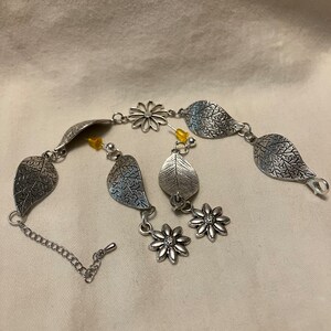 May include: A silver bracelet with a floral design and leaf charms. The bracelet has a chain closure and is decorated with small yellow beads.