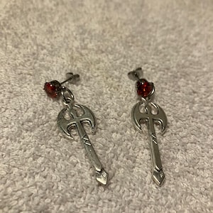 May include: A pair of silver-toned stud earrings featuring a dangling double-bladed axe charm. Each earring is accented with a small, round, faceted red gemstone set at the top of the post, connecting to the metallic axe pendant below.