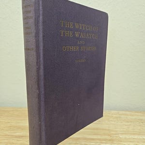 May include: A vintage, hardcover book titled "The Witch of the Wasatch and Other Stories" by Norman. The book has a dark purple cover with gold lettering. The book is standing upright on a wooden surface.