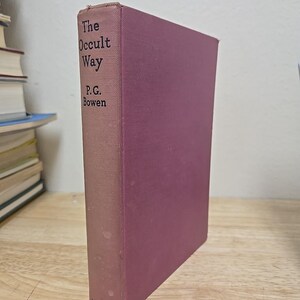 May include: A hardcover book titled "The Occult Way" by P.G. Bowen. The book has a dusty rose cover and spine with the title and author's name printed in black. The book is standing upright on a wooden surface.