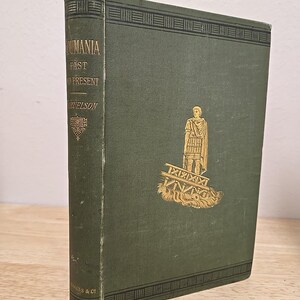 May include: A vintage hardcover book titled "Roumania Past and Present" by C. L. Samuelson. The book has a dark green cover with gold lettering and a gold illustration of a figure. The spine has the title and author's name.