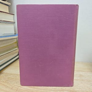 K&ouml;nnte beinhalten: Ein Hardcover-Buch mit einem einfarbigen malvenfarbenen Einband steht aufrecht auf einer Holzoberfl&auml;che. Der Buchr&uuml;cken ist sichtbar, und der Einband weist leichte Gebrauchsspuren auf. Andere B&uuml;cher sind im Hintergrund gestapelt.