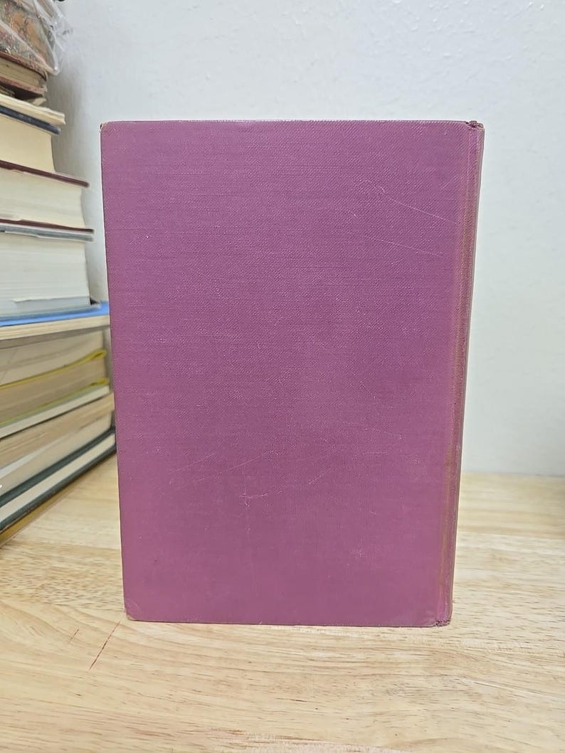 K&ouml;nnte beinhalten: Ein Hardcover-Buch mit einem einfarbigen malvenfarbenen Einband steht aufrecht auf einer Holzoberfl&auml;che. Der Buchr&uuml;cken ist sichtbar, und der Einband weist leichte Gebrauchsspuren auf. Andere B&uuml;cher sind im Hintergrund gestapelt.