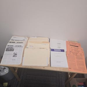 May include: A collection of documents and folders arranged on a wooden table. The items include stacks of white newsletters titled The Astrogram, several manila file folders, a white report labeled RANN, and a series of orange NASA Voyager bulletins.