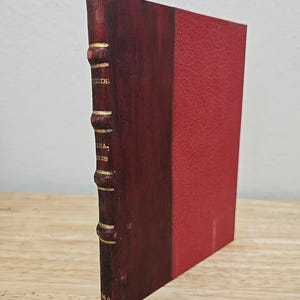 May include: A burgundy leather-bound book with gold accents on the spine. The book is closed and standing upright on a wooden surface.