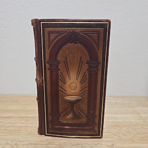 May include: A small, rectangular, brown book with a religious design. The cover features an arched doorway with columns, a chalice, and radiating light. The book has gold accents and a textured appearance, suggesting an antique or vintage style.