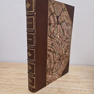 Peut inclure: Un livre ancien à couverture rigide avec un dos en cuir marron et une couverture marbrée. Le dos présente des lettres dorées en relief et des éléments décoratifs. Le livre est posé sur une surface en bois.