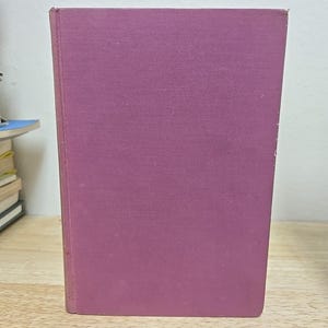 K&ouml;nnte beinhalten: Ein Hardcover-Buch mit einem einfarbigen, ged&auml;mpften lila Einband. Das Buch steht auf einer Holzoberfl&auml;che. Der Einband hat ein leicht strukturiertes Aussehen.