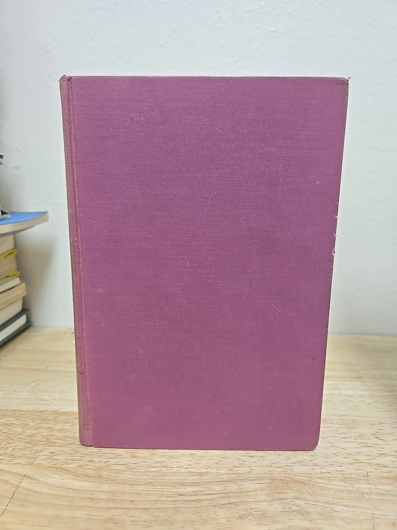 K&ouml;nnte beinhalten: Ein Hardcover-Buch mit einem einfarbigen, ged&auml;mpften lila Einband. Das Buch steht auf einer Holzoberfl&auml;che. Der Einband hat ein leicht strukturiertes Aussehen.