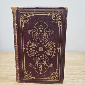 May include: A vintage, dark brown leather-bound book with gold-leaf embellishments on the cover. The design features intricate floral patterns and swirls.
