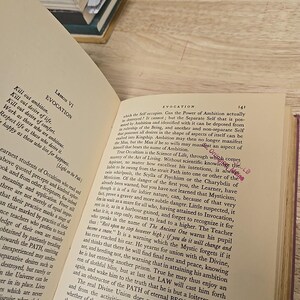 K&ouml;nnte beinhalten: Offenes Buch mit gealterten, vergilbten Seiten. Schwarzer Text f&uuml;llt die Seiten, mit der Kapitel&uuml;berschrift "Evocation" sichtbar. Der Text behandelt Okkultismus und Spiritualit&auml;t. Ein Stapel B&uuml;cher ist im Hintergrund zu sehen.