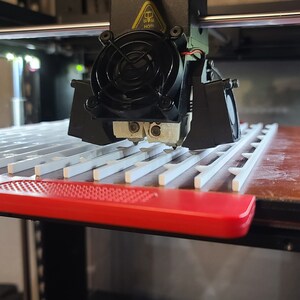 K&ouml;nnte beinhalten: Ein 3D-Drucker mit einer schwarzen D&uuml;se und einem L&uuml;fter druckt ein wei&szlig;es Plastikobjekt auf einer roten und braunen Plattform. Die Plattform hat ein Gittermuster.