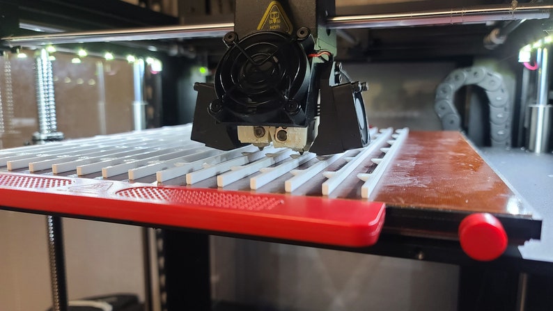 K&ouml;nnte beinhalten: Ein 3D-Drucker mit einer schwarzen D&uuml;se und einem L&uuml;fter druckt ein wei&szlig;es Plastikobjekt auf einer roten und braunen Plattform. Die Plattform hat ein Gittermuster.