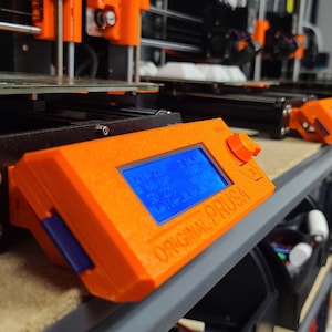 K&ouml;nnte beinhalten: Nahaufnahme eines orangefarbenen 3D-Druckersteuerpanels mit einem blauen LCD-Bildschirm, der Temperaturwerte anzeigt. Der Text "ORIGINAL PRUSA" ist auf dem Panel aufgedruckt.