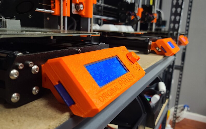K&ouml;nnte beinhalten: Nahaufnahme eines orangefarbenen 3D-Druckersteuerpanels mit einem blauen LCD-Bildschirm, der Temperaturwerte anzeigt. Der Text "ORIGINAL PRUSA" ist auf dem Panel aufgedruckt.