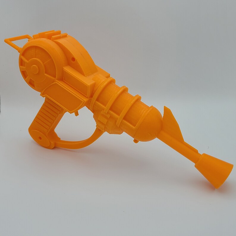 K&ouml;nnte beinhalten: Orangefarbene Spielzeugpistole aus Plastik mit einem rotierenden Rad und einem langen Lauf. Die Pistole hat einen Griff und einen Abzug.