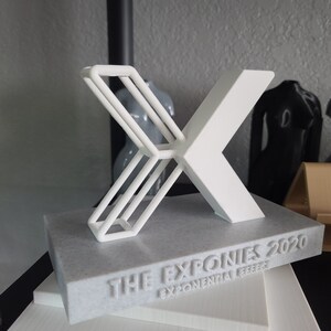 K&ouml;nnte beinhalten: Ein wei&szlig;er, 3D-gedruckter Pokal in Form eines 'X' mit dem Text "THE EXPONIES 2020 EXPONENTIAL LEAP" auf einem grauen Marmorsockel.