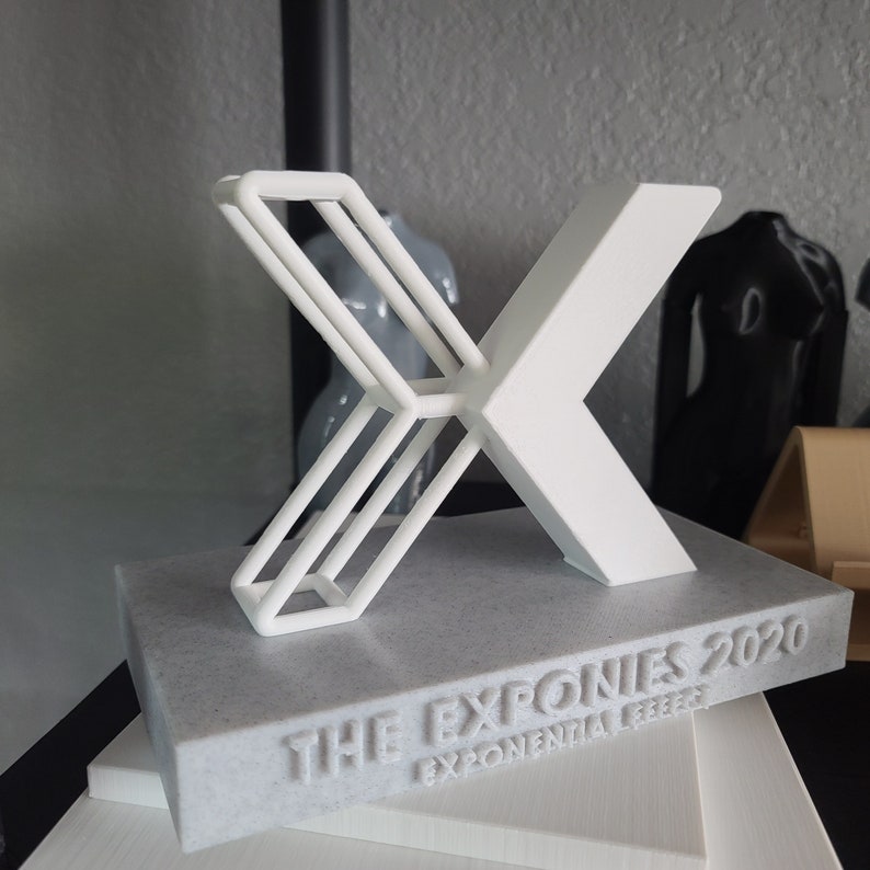 K&ouml;nnte beinhalten: Ein wei&szlig;er, 3D-gedruckter Pokal in Form eines 'X' mit dem Text "THE EXPONIES 2020 EXPONENTIAL LEAP" auf einem grauen Marmorsockel.
