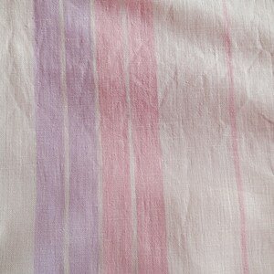 May include: A close-up of a white fabric with pink and purple stripes. The fabric has a soft, textured look.