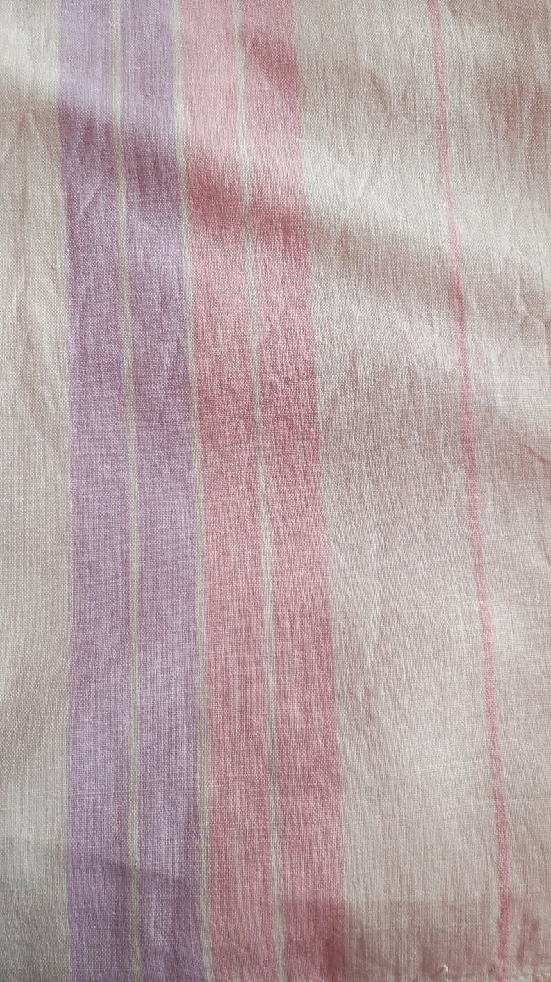 May include: A close-up of a white fabric with pink and purple stripes. The fabric has a soft, textured look.