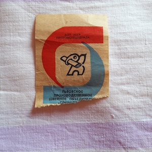 May include: A vintage label from the Lvovskoye Production Sewing Association, featuring a red and blue design with a stylised figure holding a tool. The label reads "M.L.P. USSR UKRPROMSLESHODKHALA" and "LVOVSKOE PROIZVODSTVENNOE SHVEINOE OB'EDINENIE "SPLENSO"
