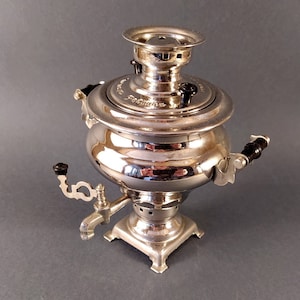 May include: A silver samovar with black accents and a spout. The samovar has a round body and a domed lid. It is a traditional Russian tea kettle.