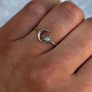 May include: A silver ring featuring a crescent moon and a sun with a turquoise stone center. The ring is delicate and minimalist, showcasing celestial-inspired jewelry. The ring is worn on a finger.