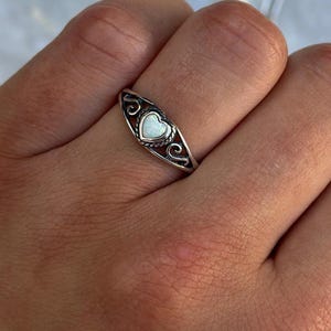 May include: A silver ring with a heart-shaped, white stone set in the center. The ring has an ornate design with swirling patterns on either side of the heart. The ring is on a person's finger.