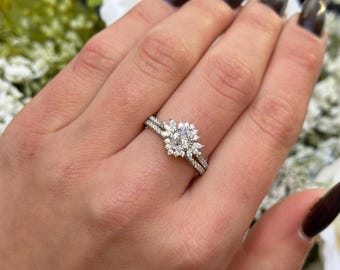 May include: A silver engagement ring set with a large oval-cut diamond center stone, surrounded by smaller accent stones in a floral design. The band is also adorned with small diamonds. The ring is on a person's finger.
