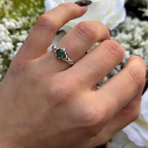 Pode incluir: Um anel de prata com uma gema verde e um pequeno detalhe de diamante. O anel é um anel Claddagh, um design tradicional irlandês.
